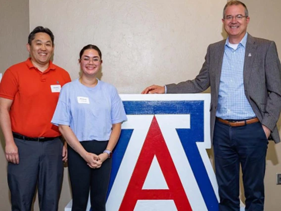 three people, smiling, standing next to UA block A logo