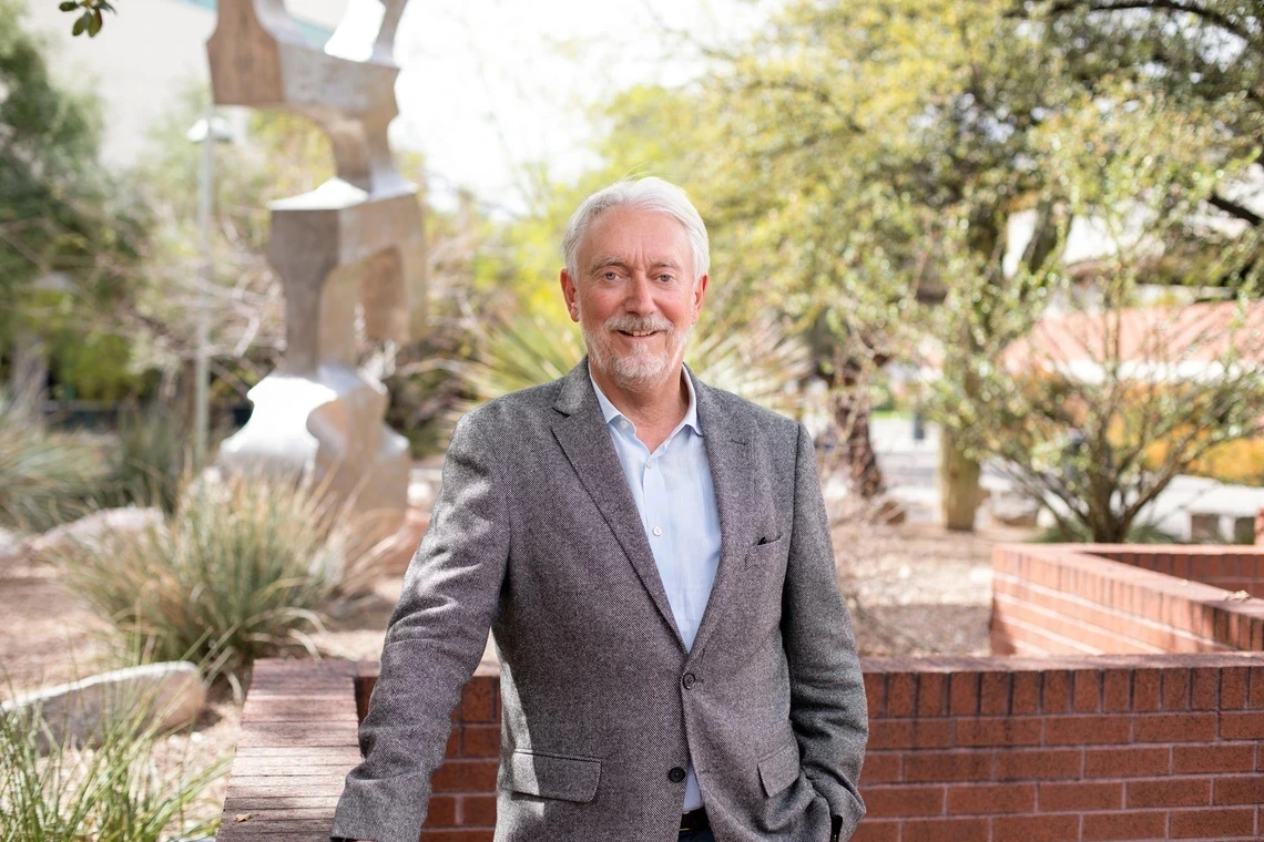 A man in a grey suit poses for a photo in front of desert plants.